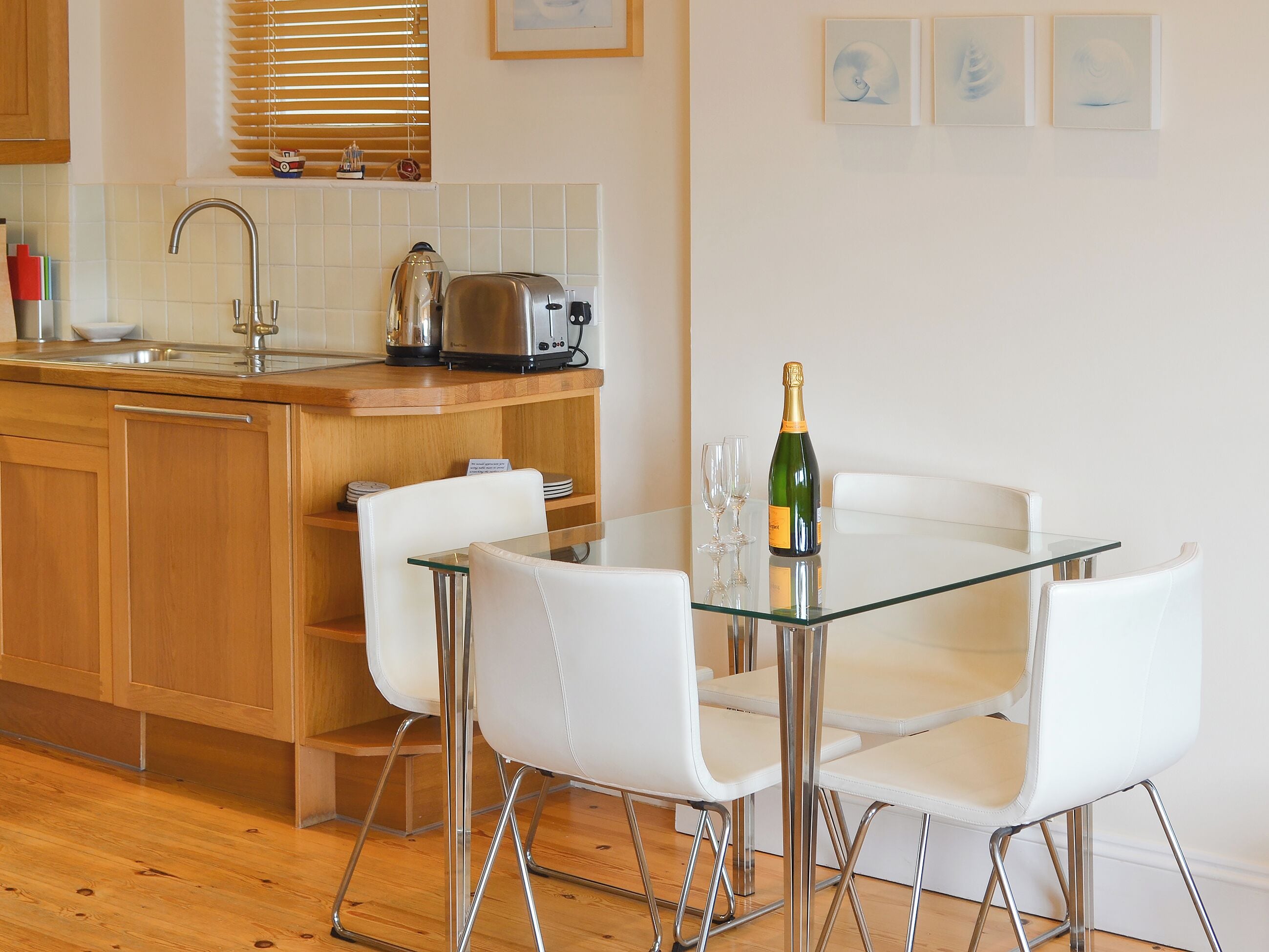 Una cocina moderna con una mesa de vidrio, sillas blancas, un tostador y una botella de champán.