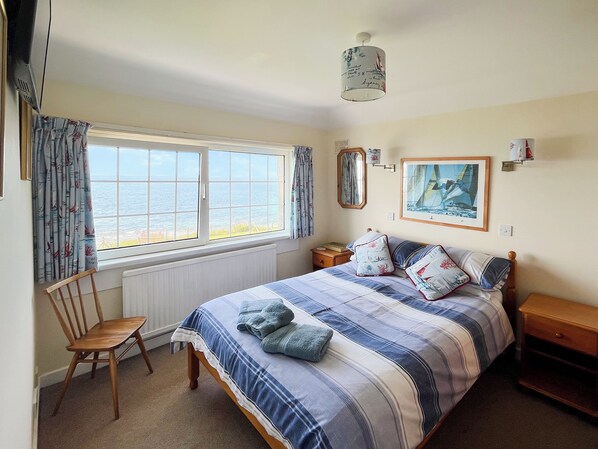 A bedroom with a bed, a chair, a nightstand, a window with curtains, a ceiling fan, and a framed picture on the wall. - Former coastguard cottage with spectacular view from Rame Head to Looe Island. (Downderry, near Looe)