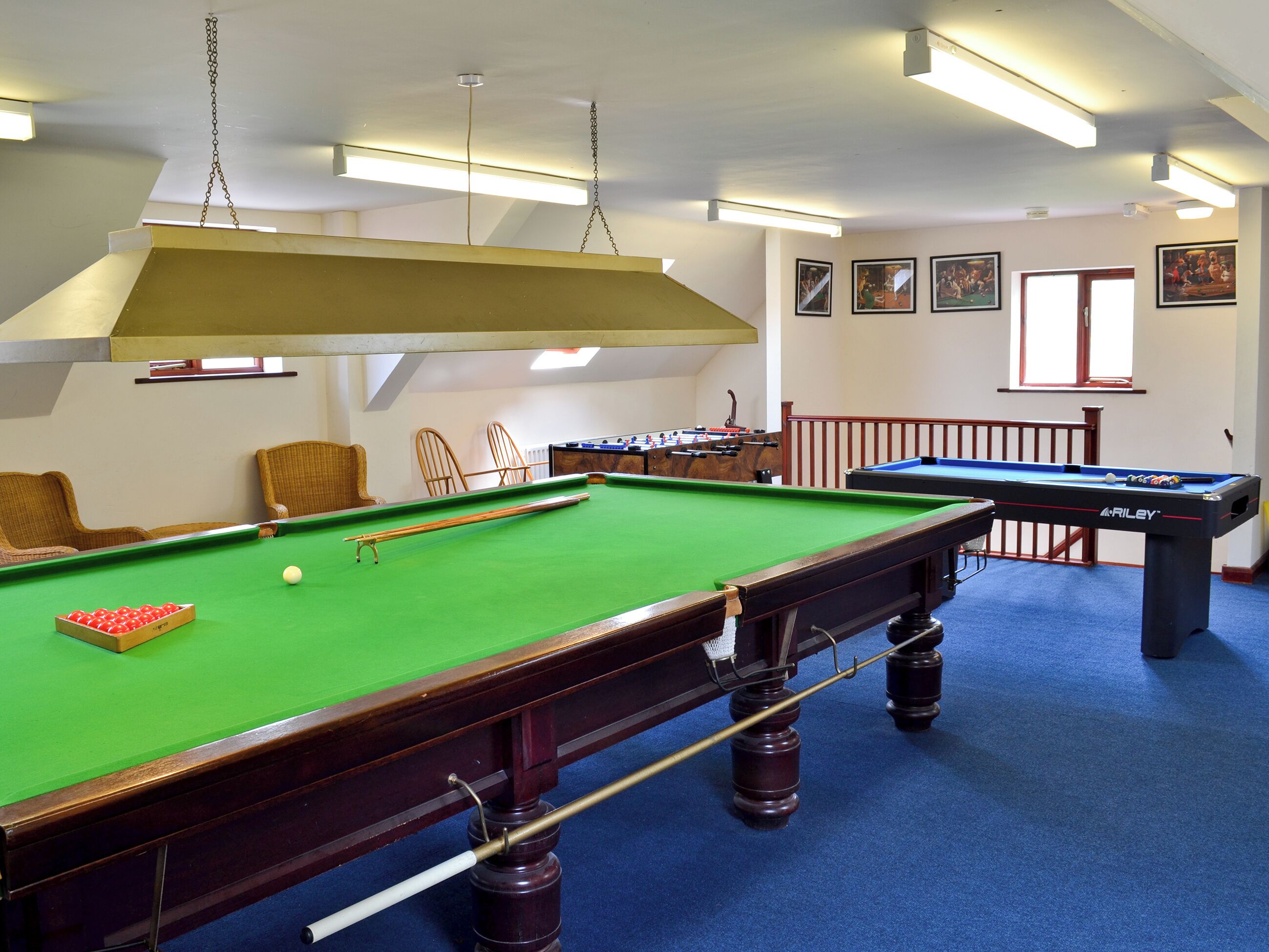 A pool table with a green felt surface, a pool cue, and a red cue ball. There are two wicker chairs and a foosball table in the background.