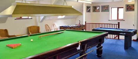 A pool table with a green felt surface, a pool cue, and a red cue ball. There are two wicker chairs and a foosball table in the background.