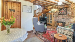 Una sala de estar acogedora con chimenea, vigas de madera, una mesa de comedor con un florero y una escalera que sube a un nivel superior.