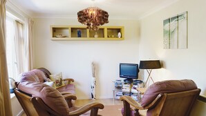 A living room with two leather armchairs, a wooden coffee table, a chandelier, a television, and a bookshelf. - 2 bedroom accommodation in Heacham (Heacham)