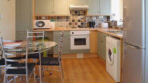 A compact kitchen with a microwave, oven, and washing machine. There is a glass table with chairs and a clock on the wall. - 2 bedroom accommodation in Heacham (Heacham)