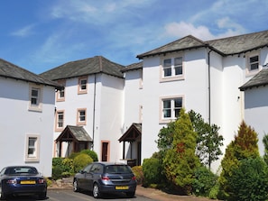 A row of white two-story houses with grey roofs, parked cars, and green bushes.