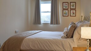 Ein ordentlich bezogenes Bett mit Kissen und Decke, ein Nachttisch mit Lampe, ein Fenster mit Vorhängen und Wandbilder mit floralem Muster.