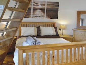 A wooden bed with a headboard, a nightstand with a lamp, a wooden dresser, and a view of the sea through a window. - 2 bedroom accommodation in Tynlon, near Rhosneigr (Tynlon, near Rhosneigr)