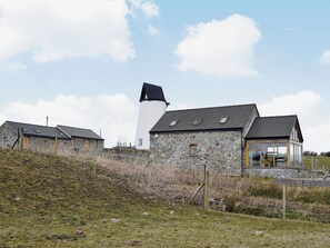 Un bâtiment en pierre surmonté d’une tour blanche, une structure en bois dotée d’une paroi vitrée, et un mur de pierre avec une clôture.