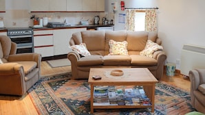 A cozy living room with a sofa, armchairs, and a coffee table with magazines. A kitchen with cabinets and appliances is visible in the background. - 3 bedroom accommodation in John O’ Groats (John O’ Groats)