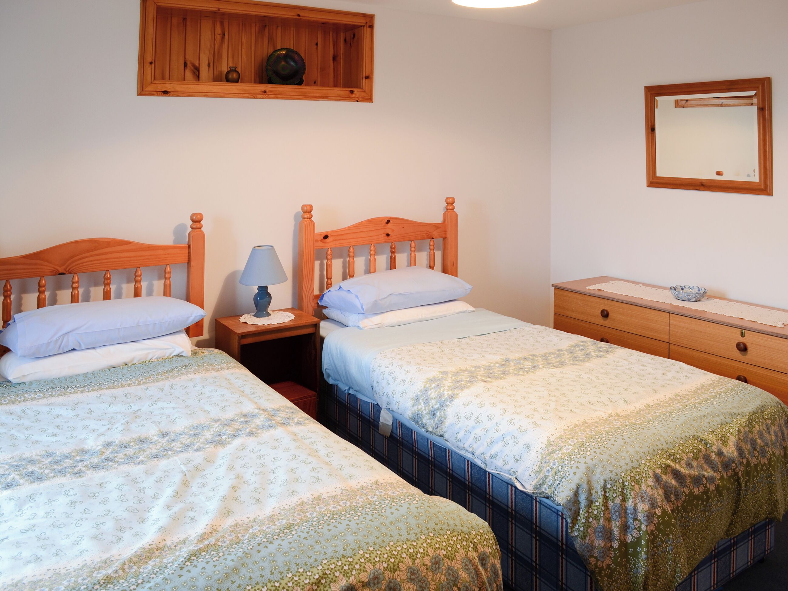 Un dormitorio con dos camas de madera, una mesita de noche con lámpara, un aparador, un espejo y una estantería con adornos.