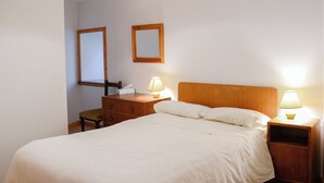 A neatly made bed with white bedding, a wooden nightstand with a lamp, a wooden dresser, and two framed mirrors on the wall. - 3 bedroom accommodation in John O’ Groats (John O’ Groats)