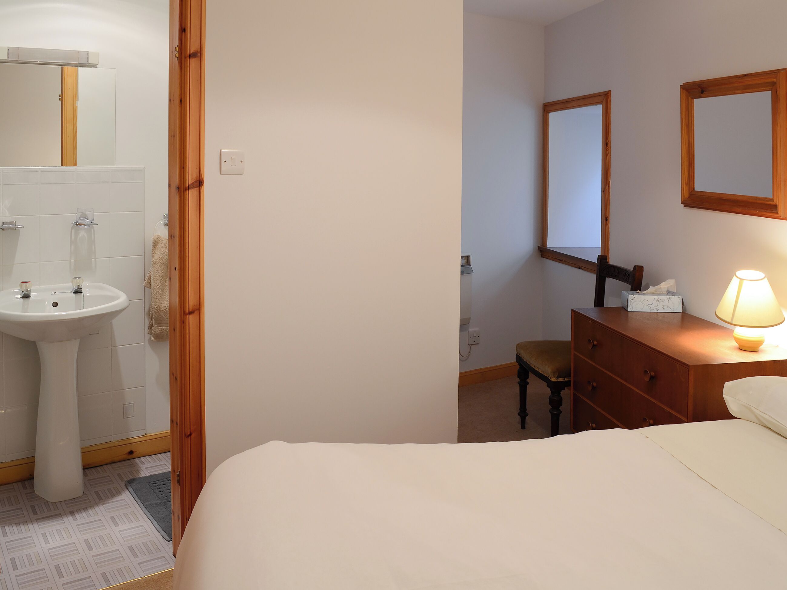 A bedroom with a bed, a bedside table with a lamp, a wooden dresser, a chair, and a bathroom with a sink and mirror.