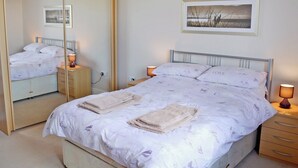 A neatly made bed with a patterned bedspread, two bedside lamps, a bedside table, a mirror, and a framed picture on the wall. - 2 bedroom accommodation in Towan Cross, near Porthowan (Towan Cross, near Porthowan)