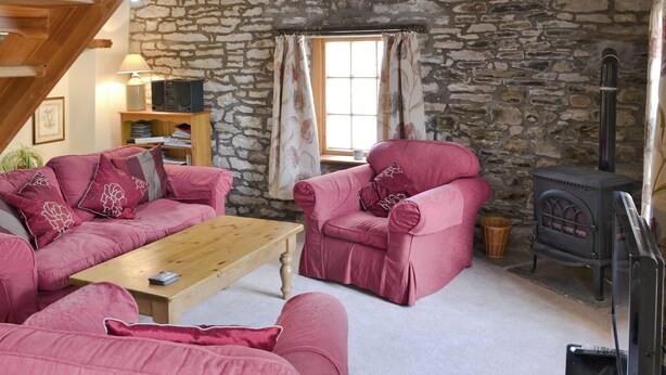 Un salon chaleureux avec un mur en pierre, un plafond en bois, une cheminée et des meubles recouverts d’un tissu rose.