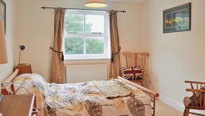 Un dormitorio con cama, una silla, escritorio, ventana con cortinas y un cuadro en la pared.