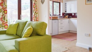 Un sofá verde con cojines amarillos en una habitación con área de cocina, incluyendo armarios blancos y una ventana con cortinas florales.