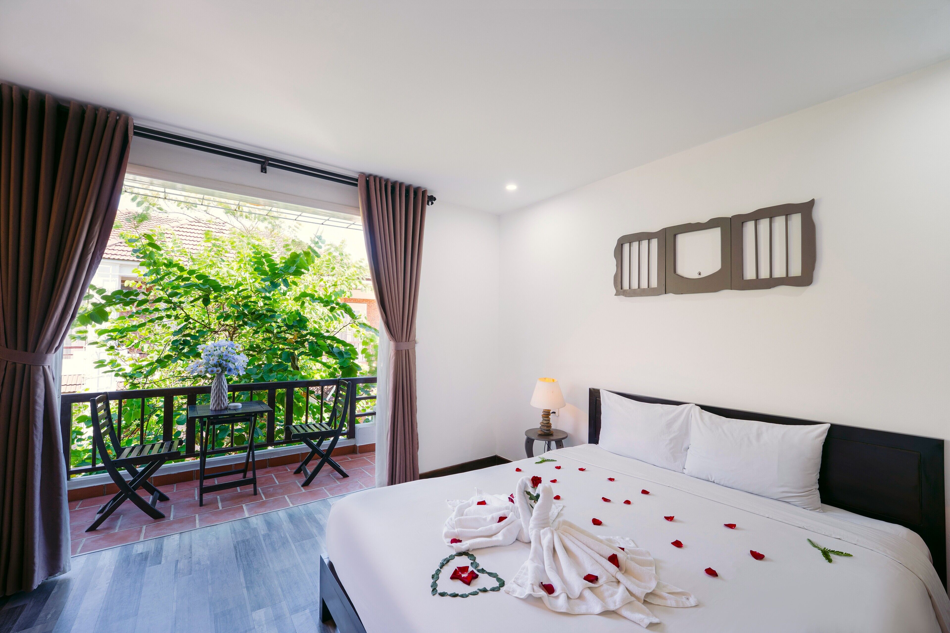 A hotel room with a bed, a small table with a vase, a chair, and a balcony with a view of greenery.