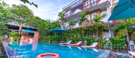 Una piscina con sillas de descanso y un flotador rojo, rodeada de abundante vegetación y un edificio de varios pisos con balcones.