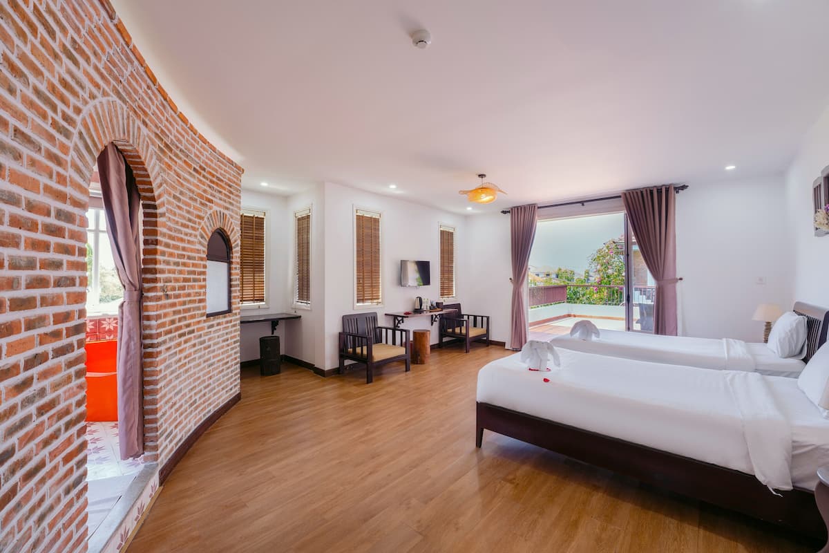 A hotel room with a brick wall, a desk with a chair, a bed, and a balcony with curtains.