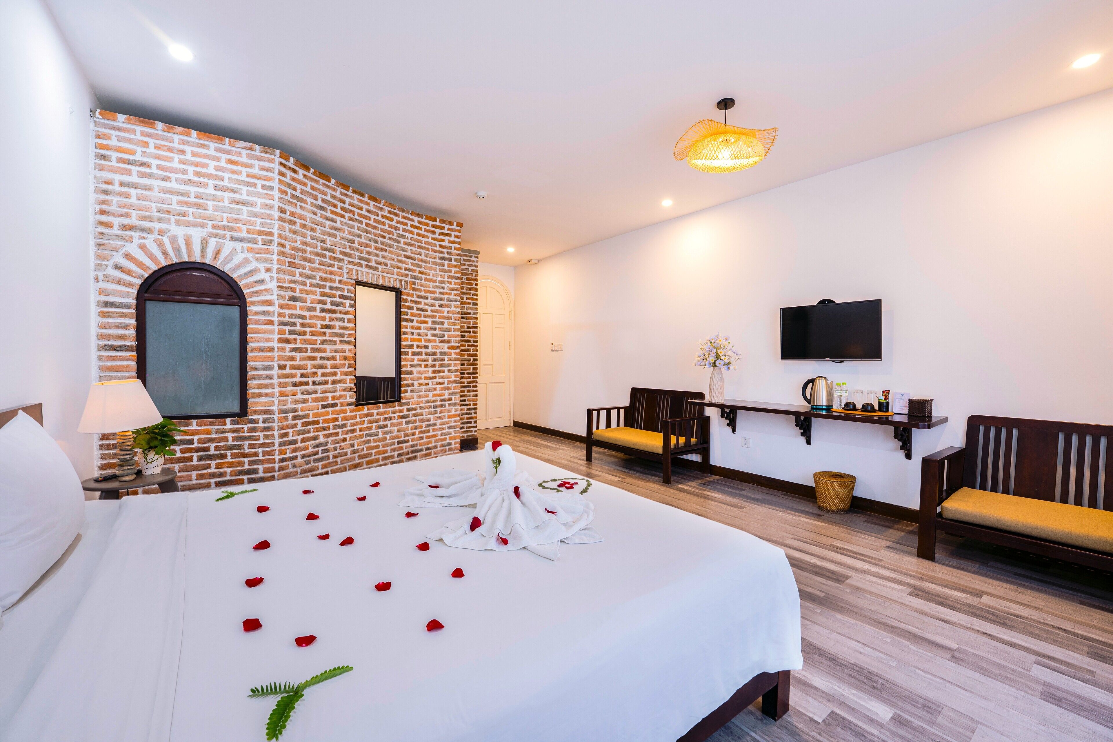 Una habitación de hotel con pared de ladrillo decorativa, una cama con pétalos de rosa y follaje, un televisor y una zona de estar con sillas y una mesa.
