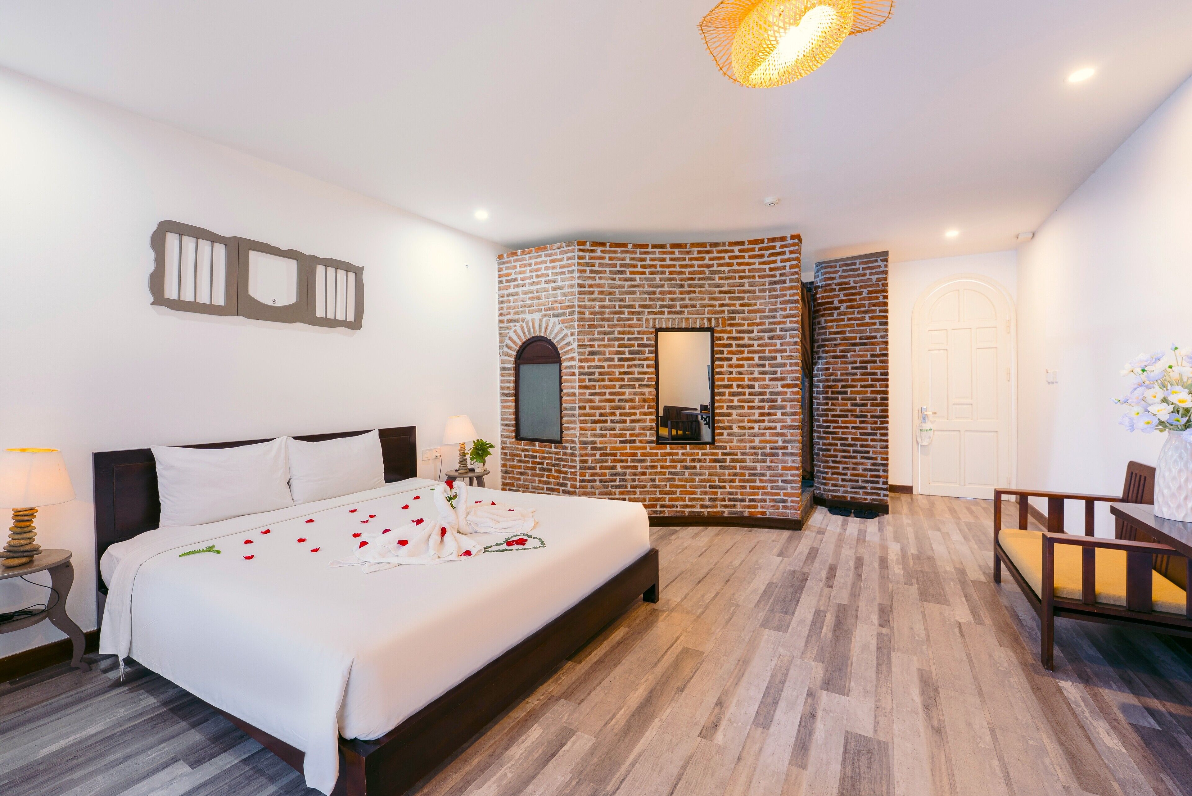 A hotel room with a brick accent wall, a bed with rose petals, a wooden floor, a desk with a chair, and a hanging light fixture.
