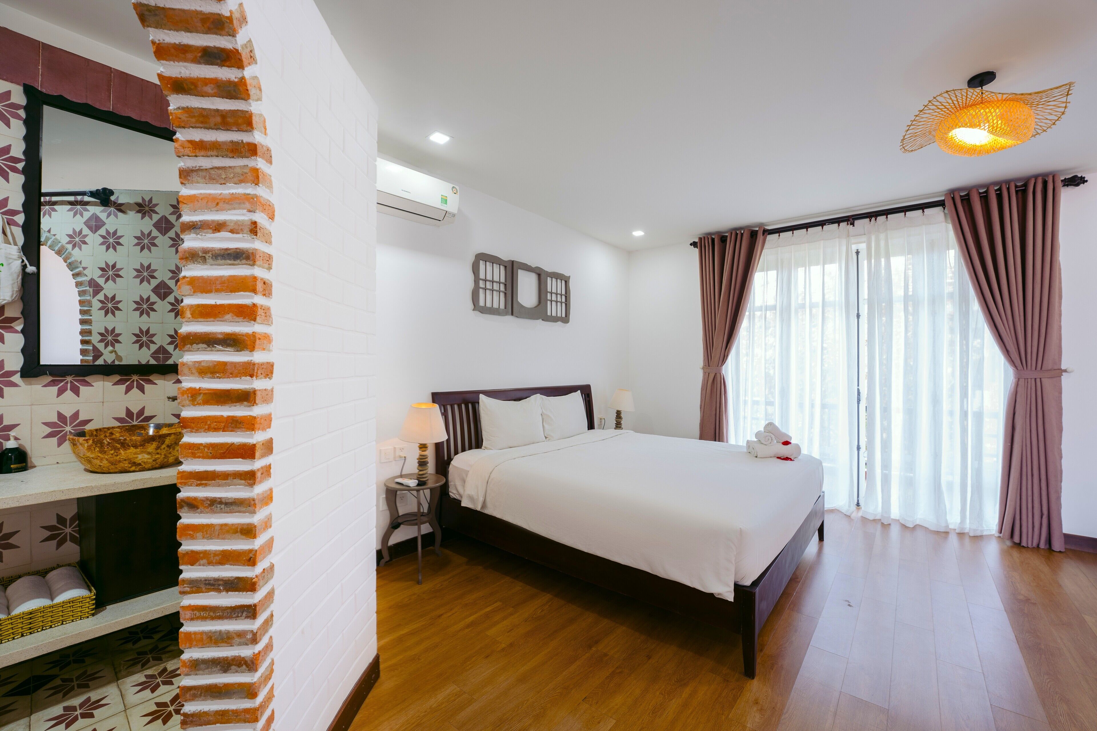 Une chambre d’hôtel avec un plancher en bois, un lit avec une literie blanche, une table de chevet avec une lampe, un climatiseur fixé au mur et une fenêtre avec des rideaux.