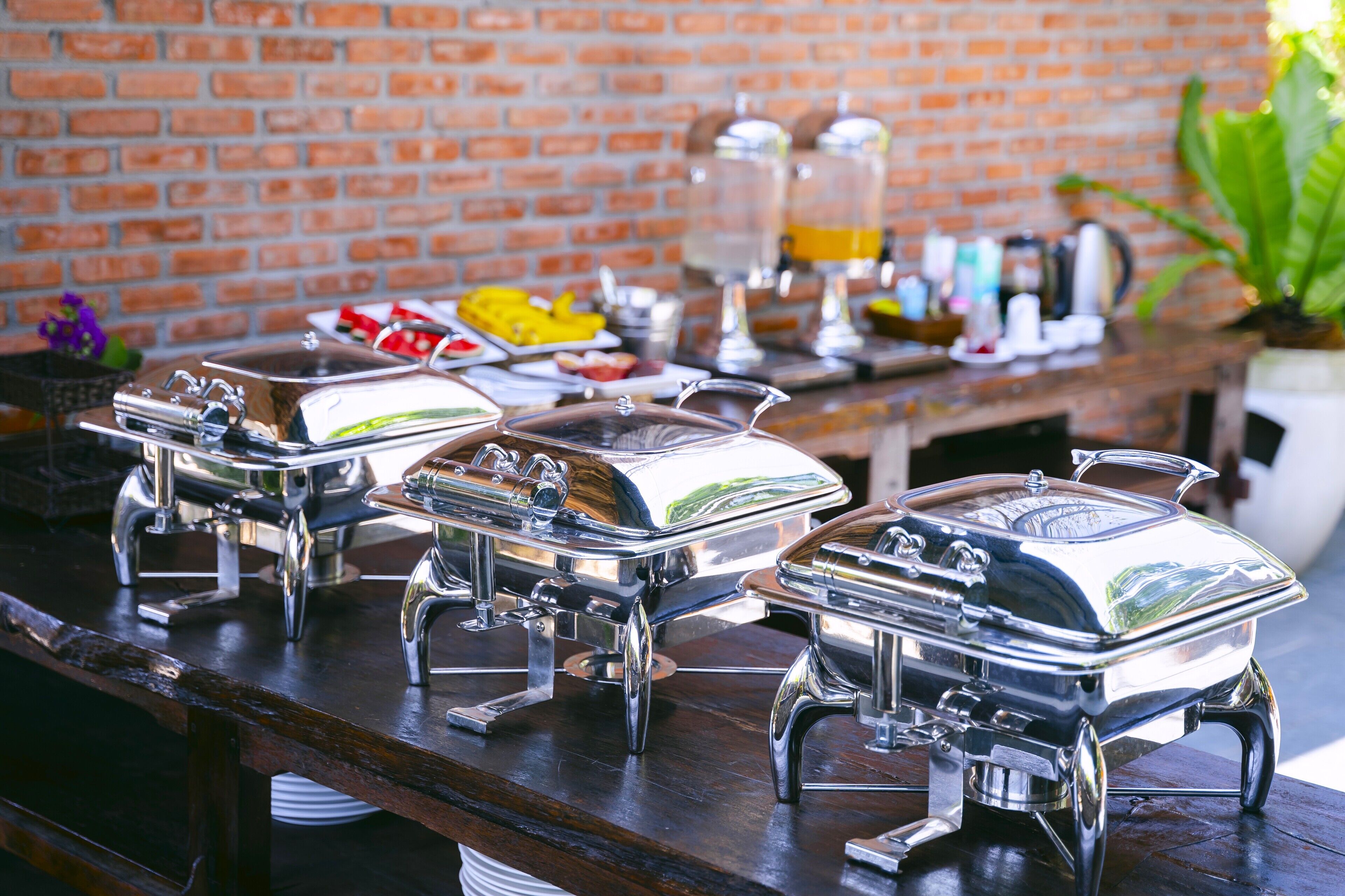 A buffet setup with multiple stainless steel chafing dishes on a wooden table against a brick wall.