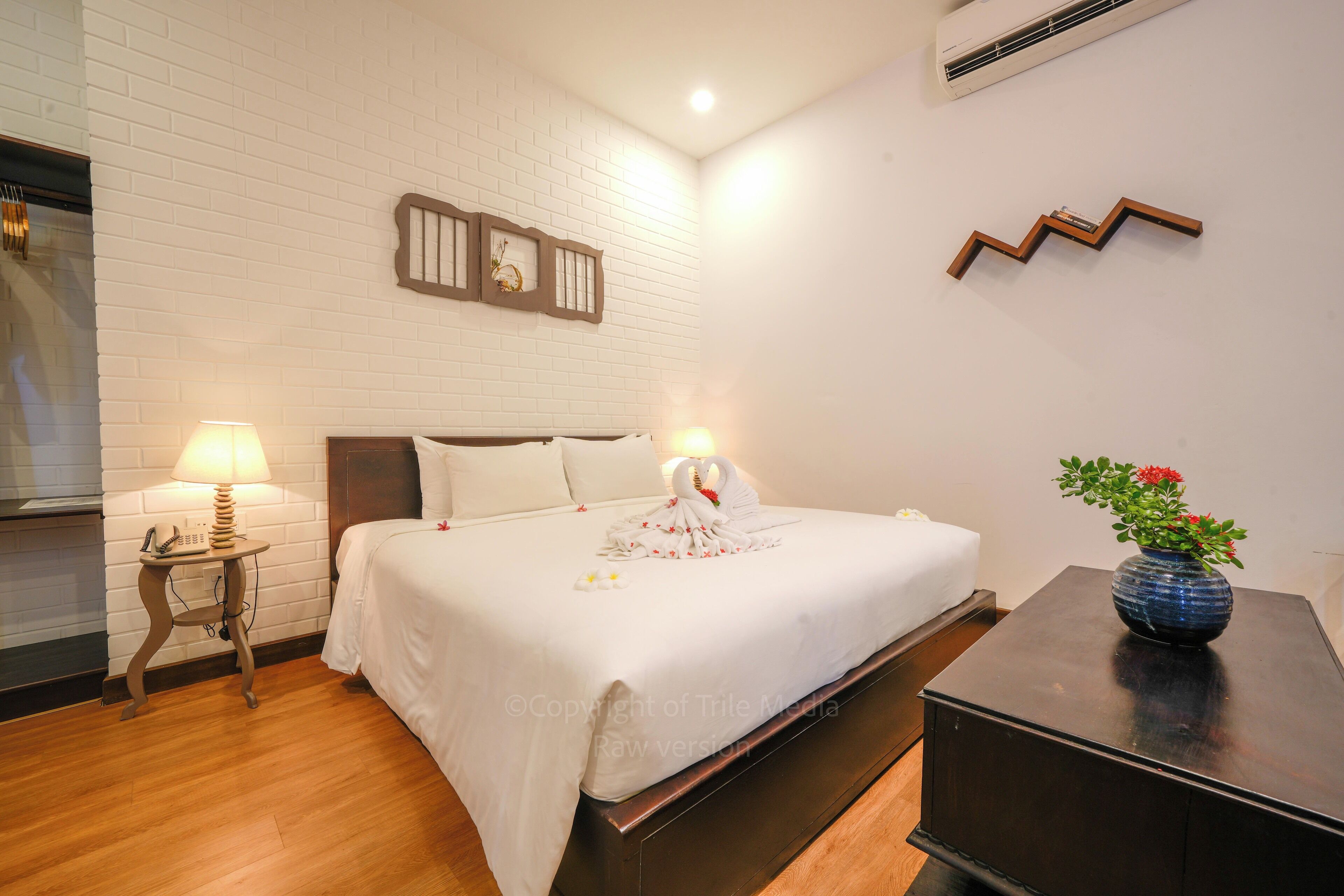 A neatly made bed with white bedding, a bedside lamp, a small table with a vase of flowers, and a wall-mounted air conditioner.