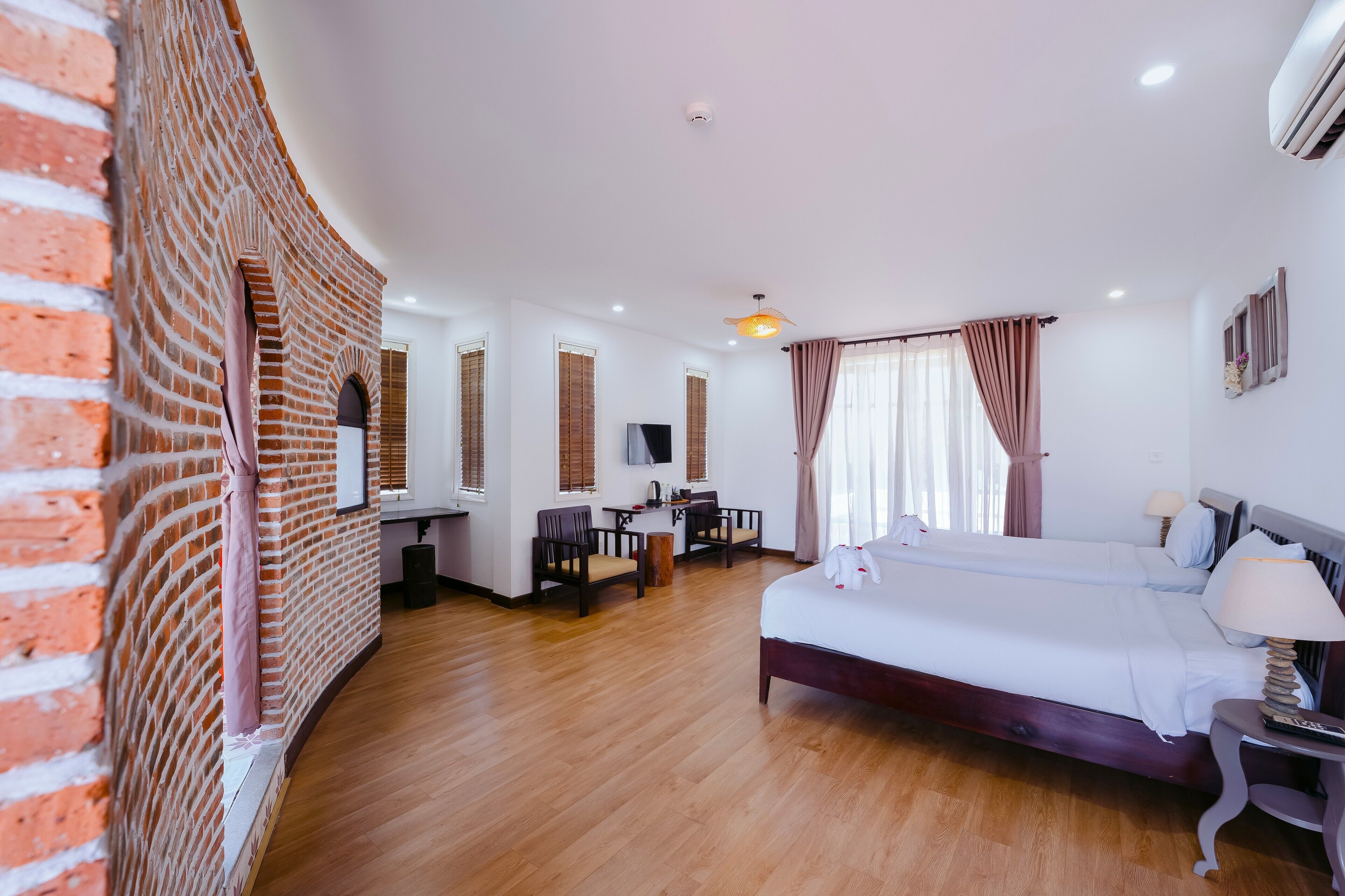 A hotel room with a brick accent wall, a wooden floor, a bed with white linens, a desk with a chair, a TV, and a window with curtains.