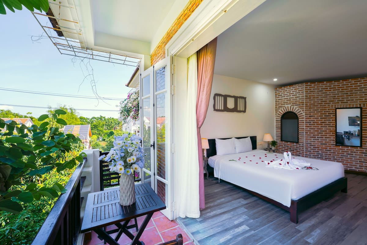 A bedroom with a bed, a nightstand, a window with curtains, a brick wall, and a balcony with a table and flowers.