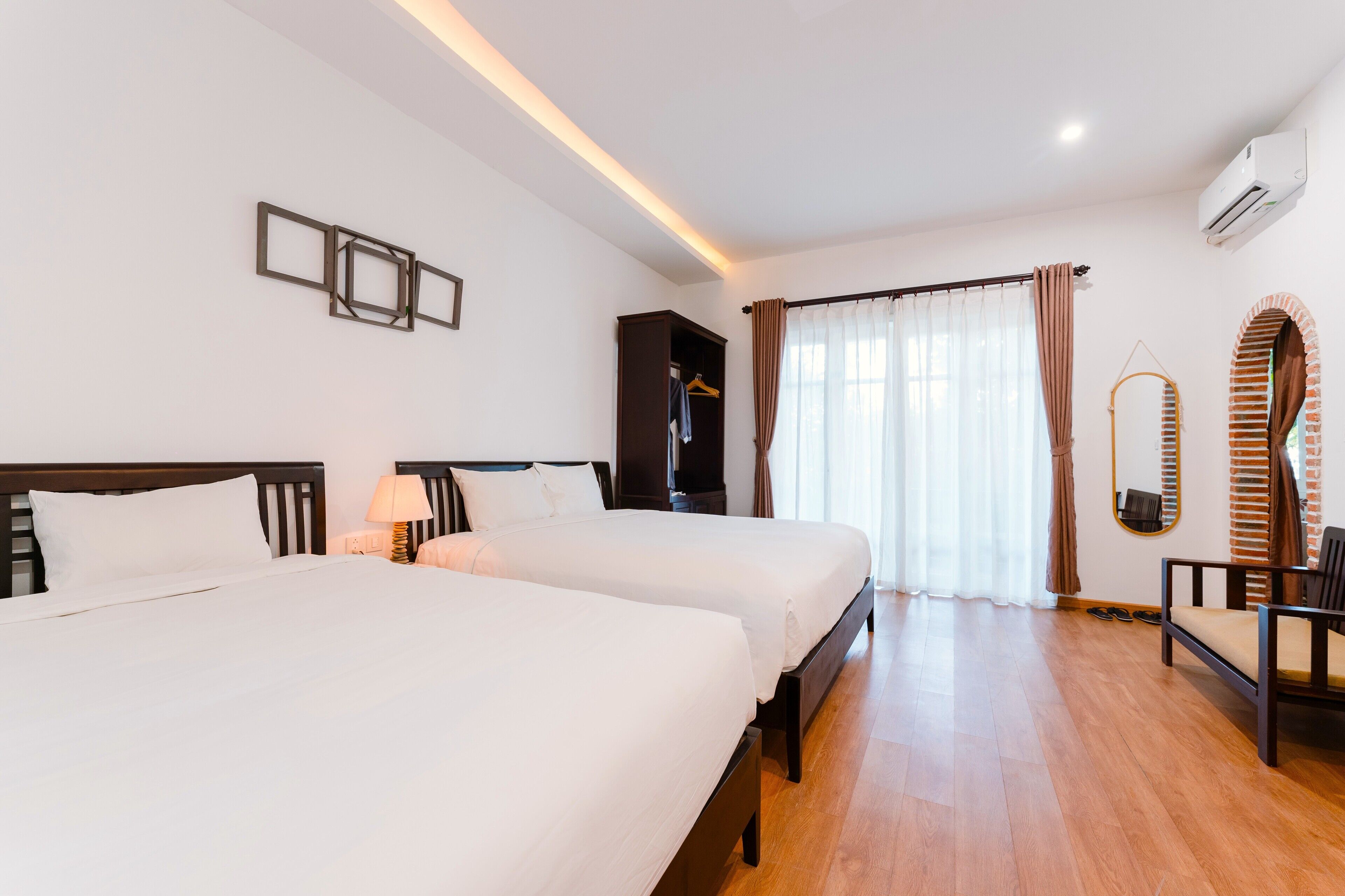 A hotel room with two beds, a wardrobe, a desk with a chair, and a large window with curtains.