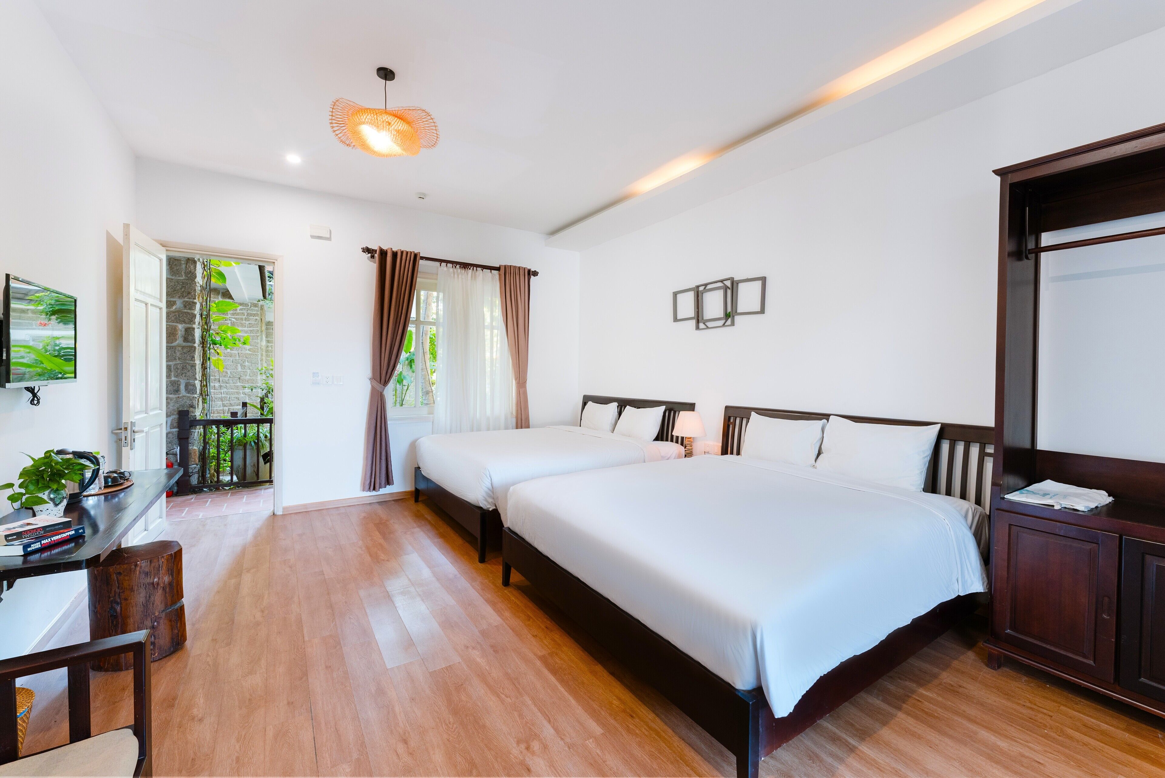 A hotel room with two beds, a desk with a chair, a TV, and a balcony with plants.