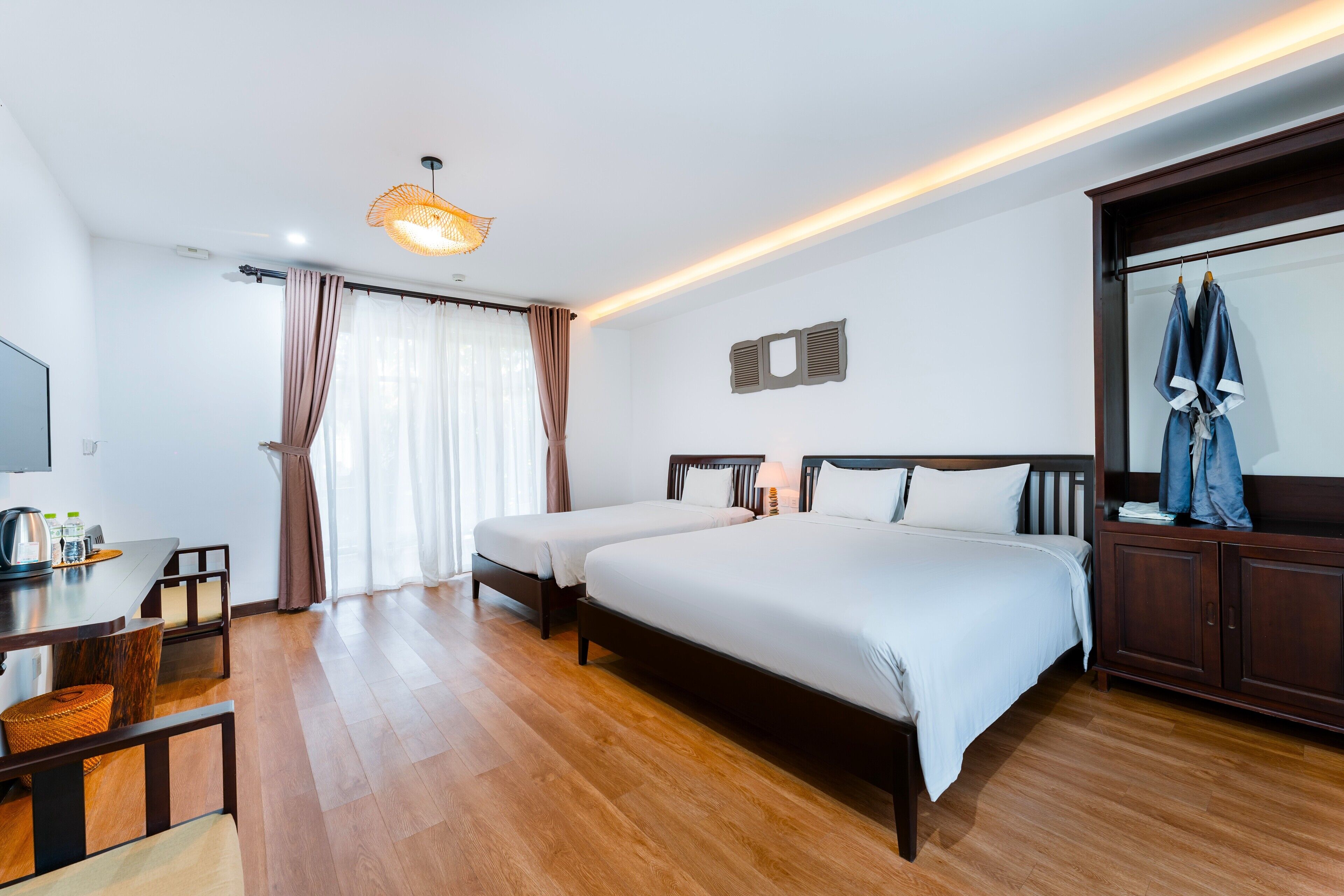 A hotel room with two beds, a wooden floor, a desk with a chair, a television, and a wardrobe.