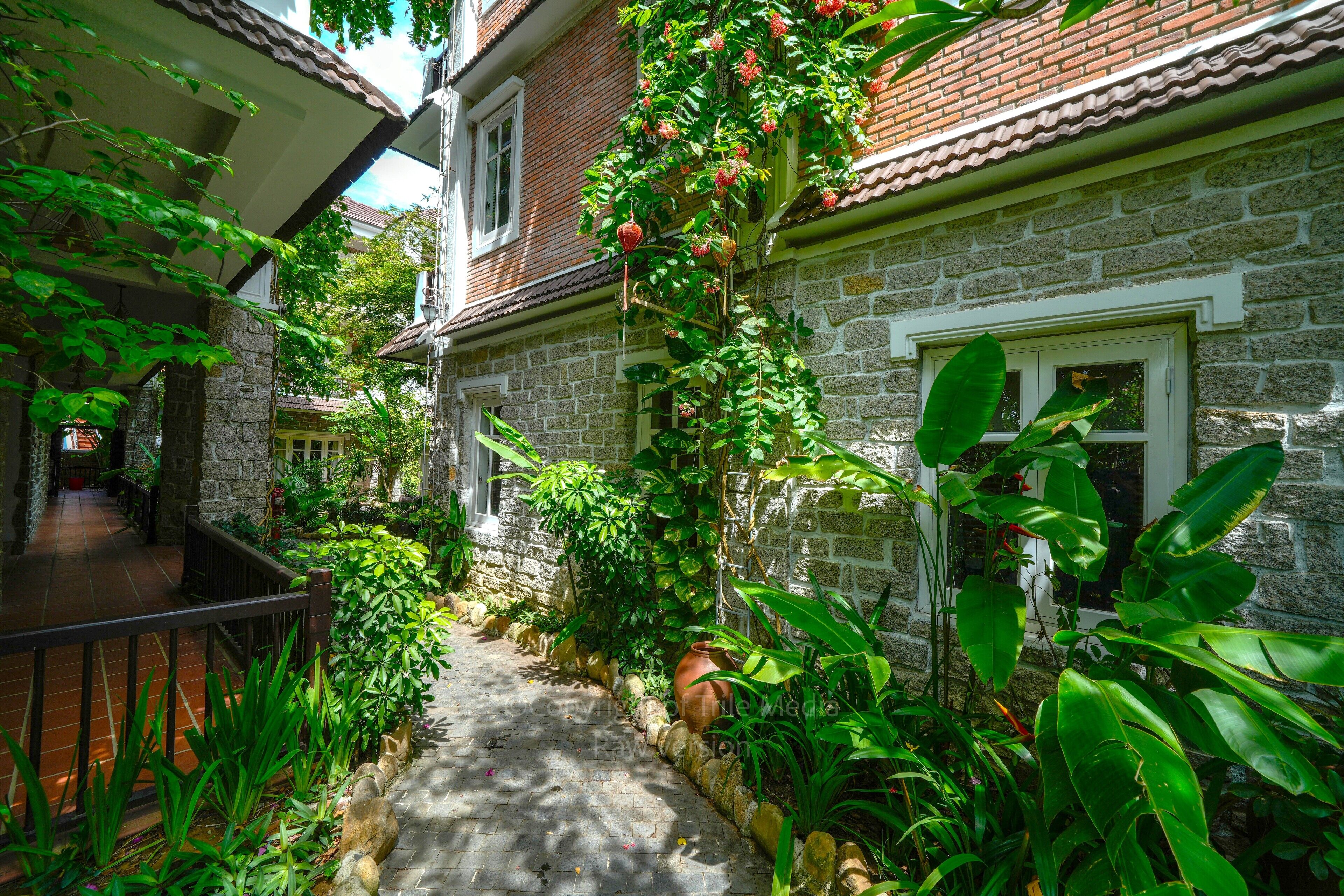 A narrow stone pathway flanked by lush greenery and a brick building with a tiled roof.