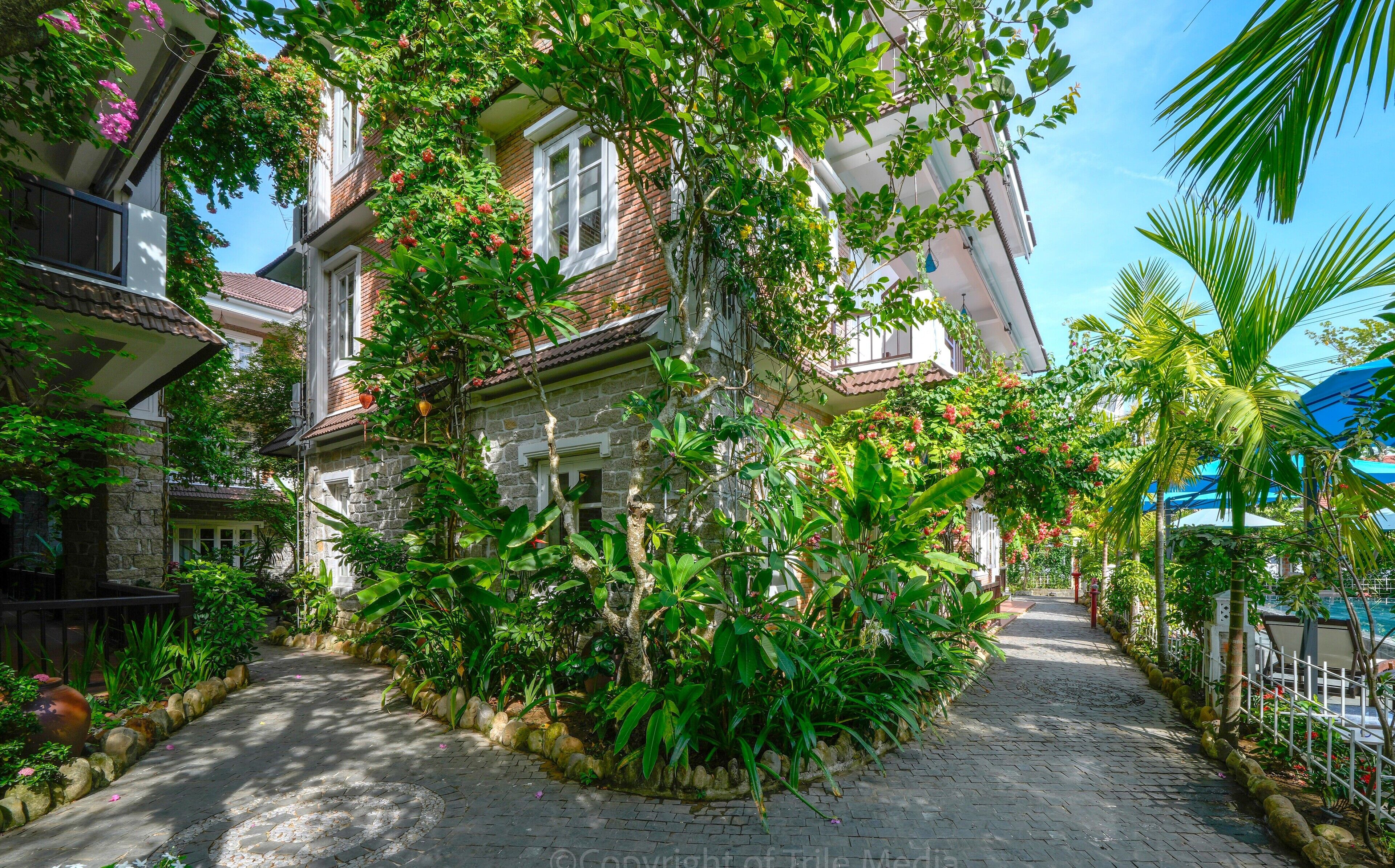 A narrow cobblestone pathway flanked by lush greenery and traditional buildings with stone walls and tiled roofs.