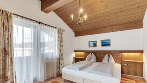A bedroom with a wooden ceiling, a bed with white bedding, a nightstand, and a view of a landscape through sheer curtains. - Apartmenthaus Jagdhof (Reith bei Seefeld)
