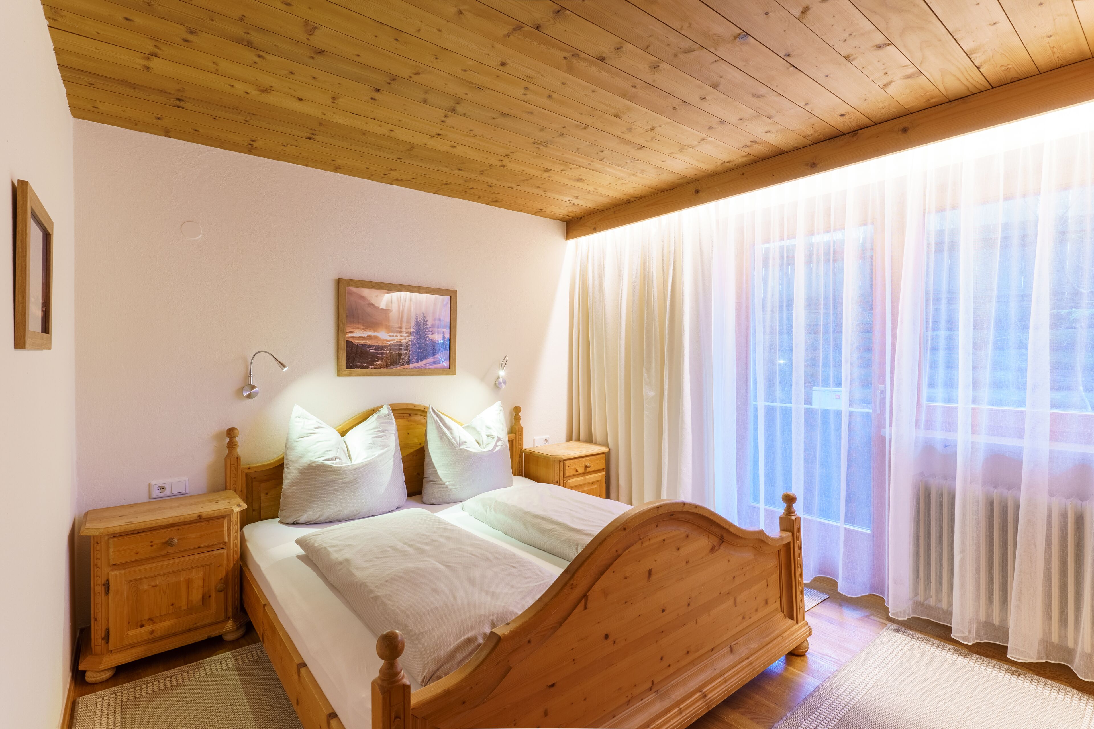 Una cama de madera con cabecera, una mesita de noche y una ventana con cortinas traslúcidas.