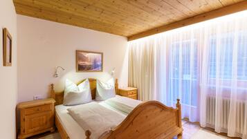 Una cama de madera con cabecera, una mesita de noche y una ventana con cortinas traslúcidas.