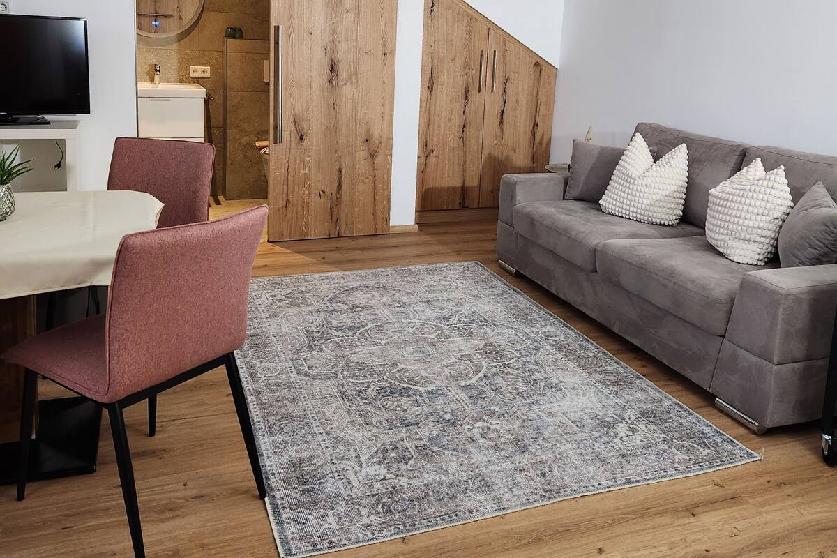 A modern living room with a grey sofa, a dining area with a table and chairs, and a large wooden wardrobe.