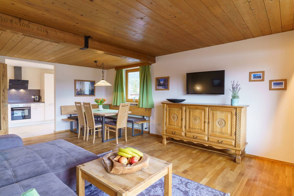 A modern living room with a wooden ceiling, a sofa, a dining table with chairs, a TV, and a wooden cabinet.