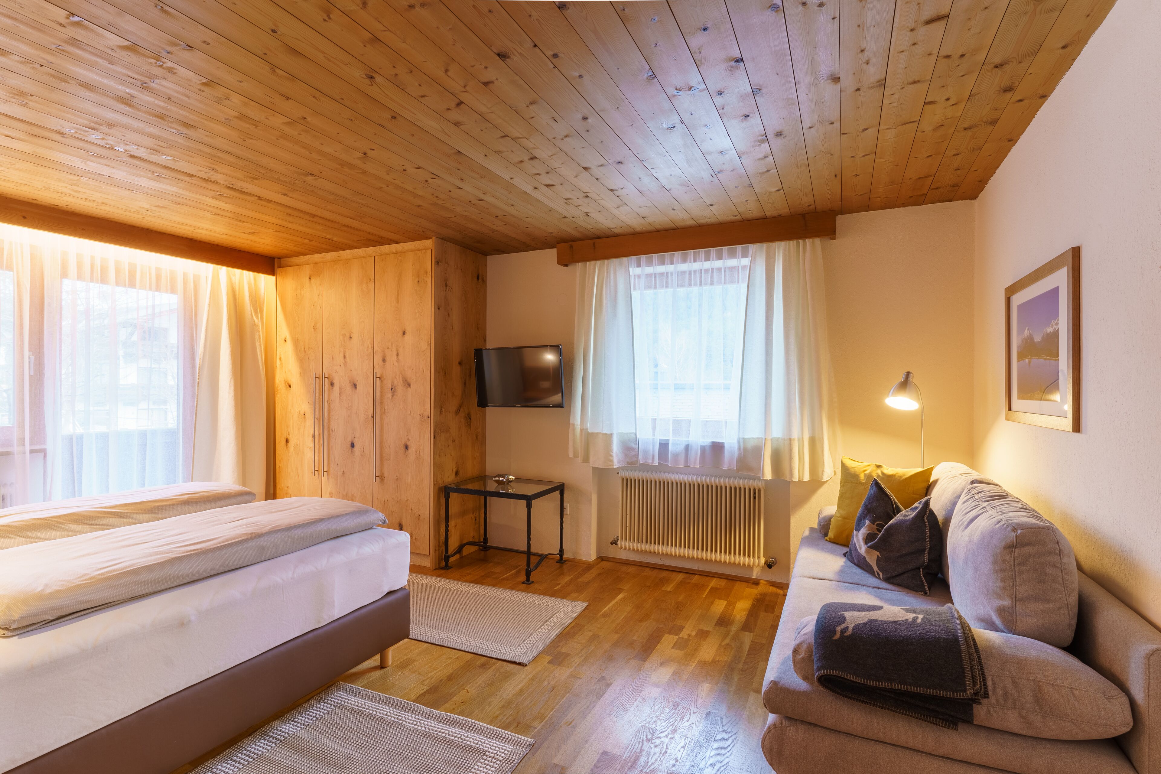 Ein gemütliches Zimmer mit Holzbalkendecke, einem Bett, einer Couch, einem Fernseher und einem Fenster mit Vorhängen.