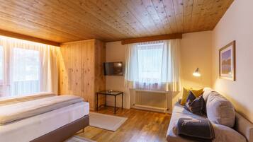 Ein gemütliches Zimmer mit Holzbalkendecke, einem Bett, einer Couch, einem Fernseher und einem Fenster mit Vorhängen.