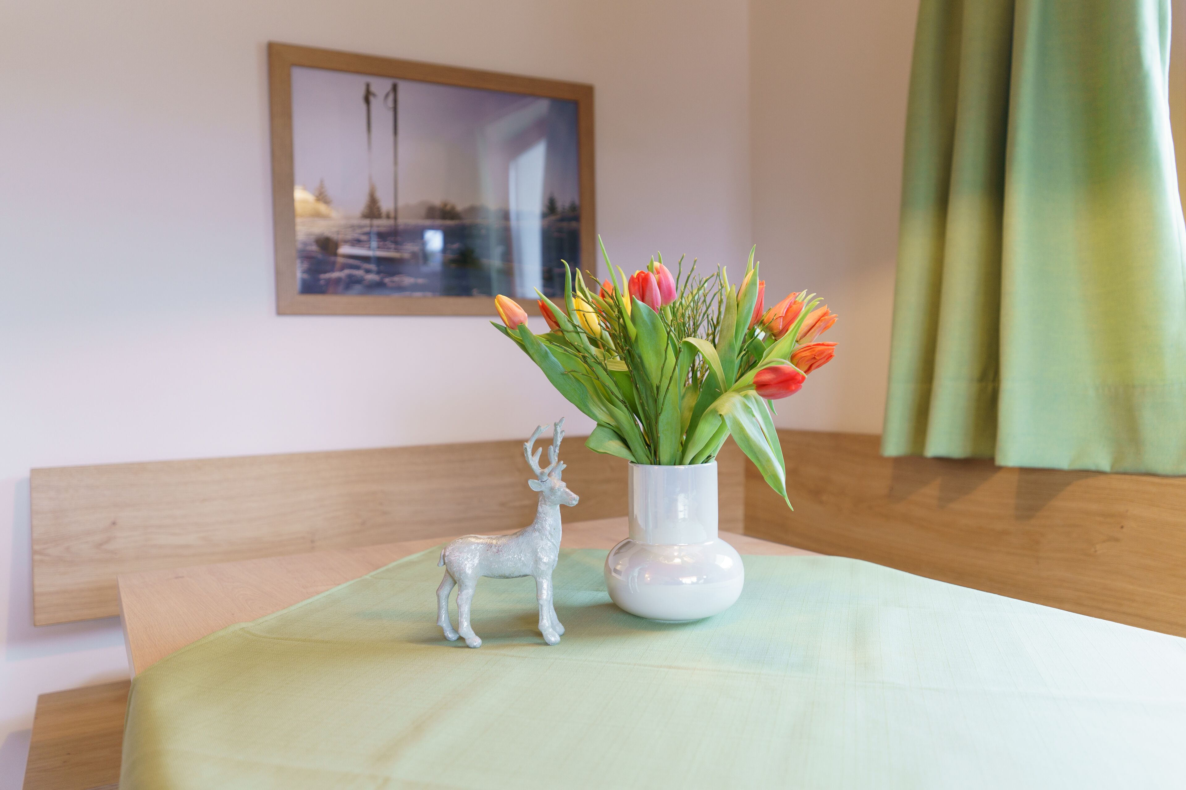 Ein Tisch mit einer weißen Vase, die rote und gelbe Tulpen enthält, einer silbernen Hirschfigur und einem gerahmten Bild an der Wand.