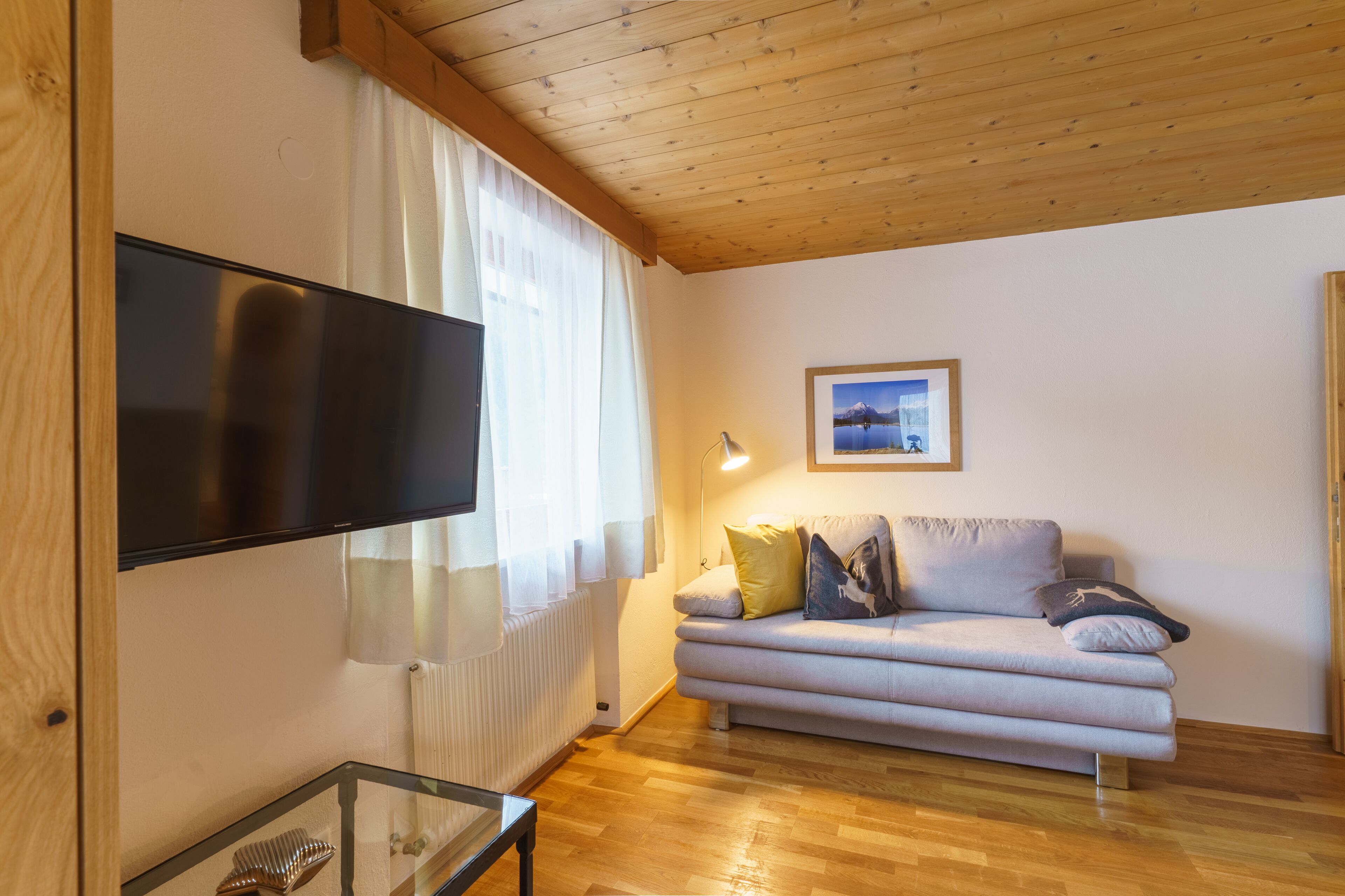 A living room with a flat-screen TV, a sofa with cushions, a framed picture on the wall, and a wooden ceiling.