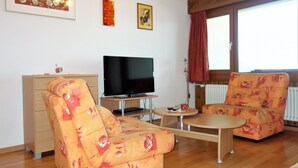 Ein Wohnzimmer mit einer Couch, einem Fernseher, einem kleinen Tisch und einem Fenster mit Vorhängen.