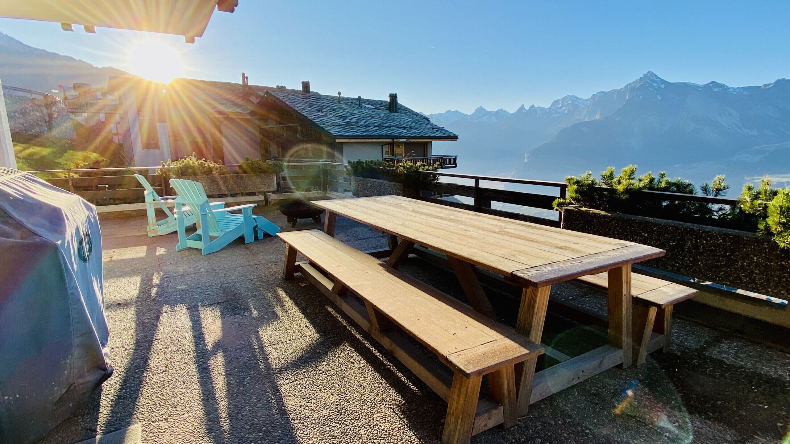 Een terras buiten met houten banken en stoelen, een barbecue en uitzicht op bergen.