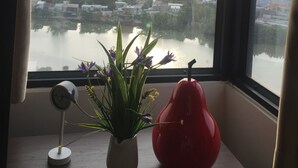 A view of a cityscape through a window with curtains, a clock, and a vase with flowers on a windowsill.