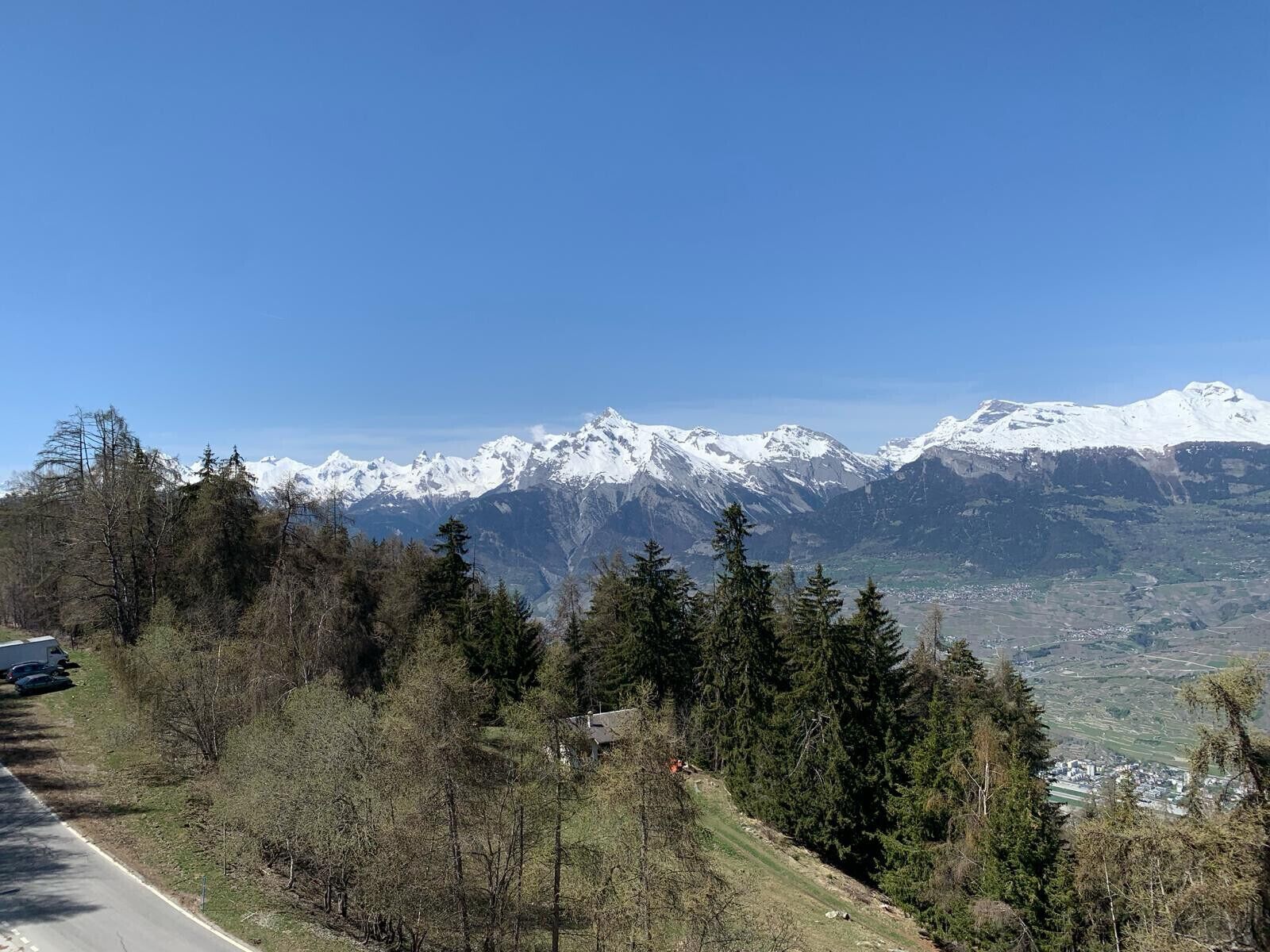 A mountainous landscape with snow-capped peaks, a road with parked cars, and dense forest in the foreground.