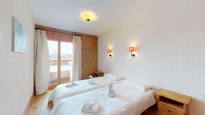 A hotel room with two beds, a wooden headboard, a balcony with a view of snow-capped mountains, and two wall-mounted lamps.