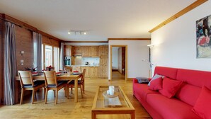 A spacious living area with a red sofa, wooden dining table, and chairs. A kitchen with wooden cabinets and a modern design is visible in the background.