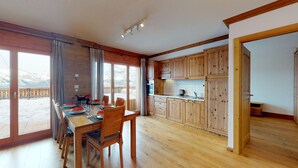 A wooden dining table with chairs, a kitchen with wooden cabinets, and a view of snow-covered mountains through large windows.